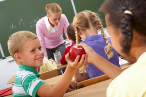 Portrait of schoolgirl passing red apple to classmate during lesson